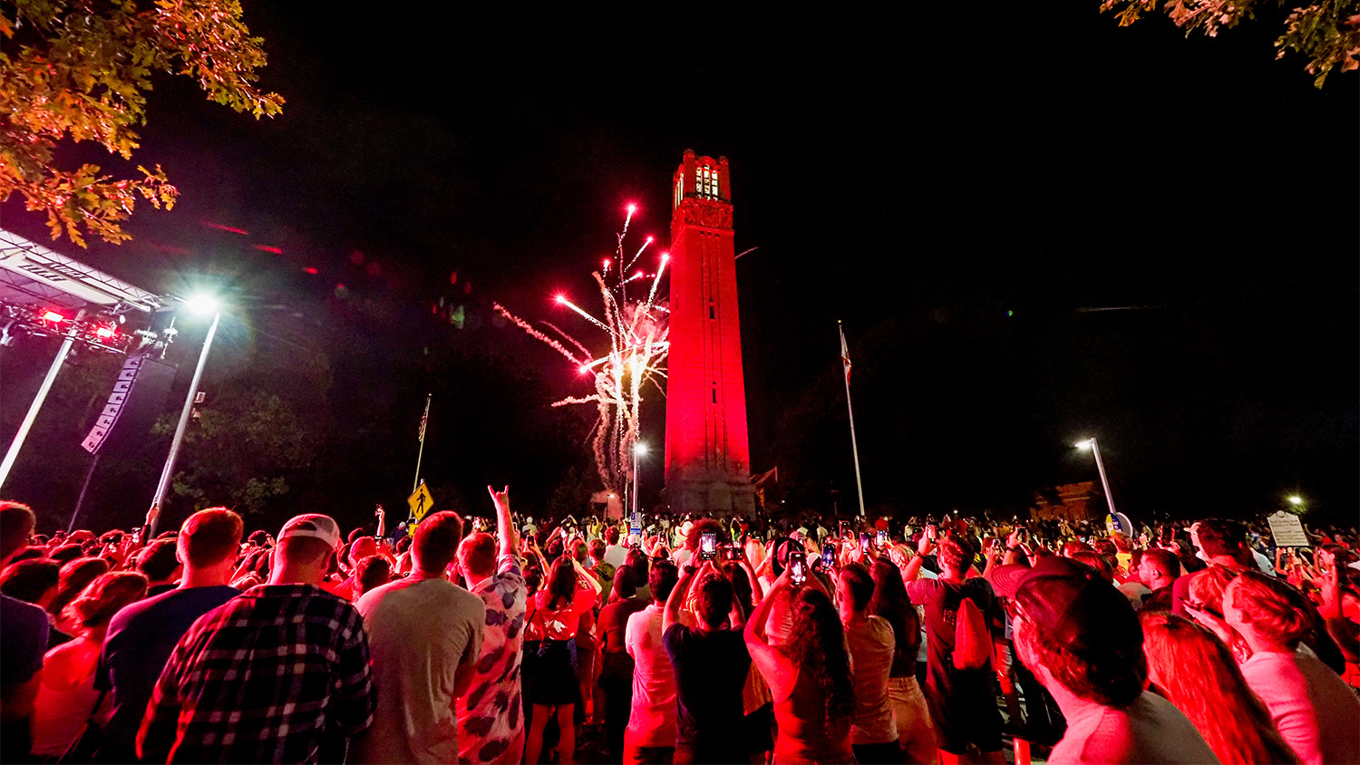 Belltower lit in red at night with fireworks in background and cheering crowd in foreground.